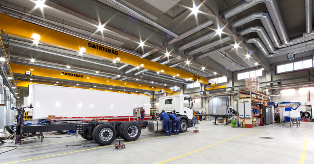 Tilava teollisuusvarasto korkealla katolla ja nostureilla. Työntekijät sinisissä univormuissa korjaavat tai kokoavat suurta valkoista kuorma-auton perävaunua.