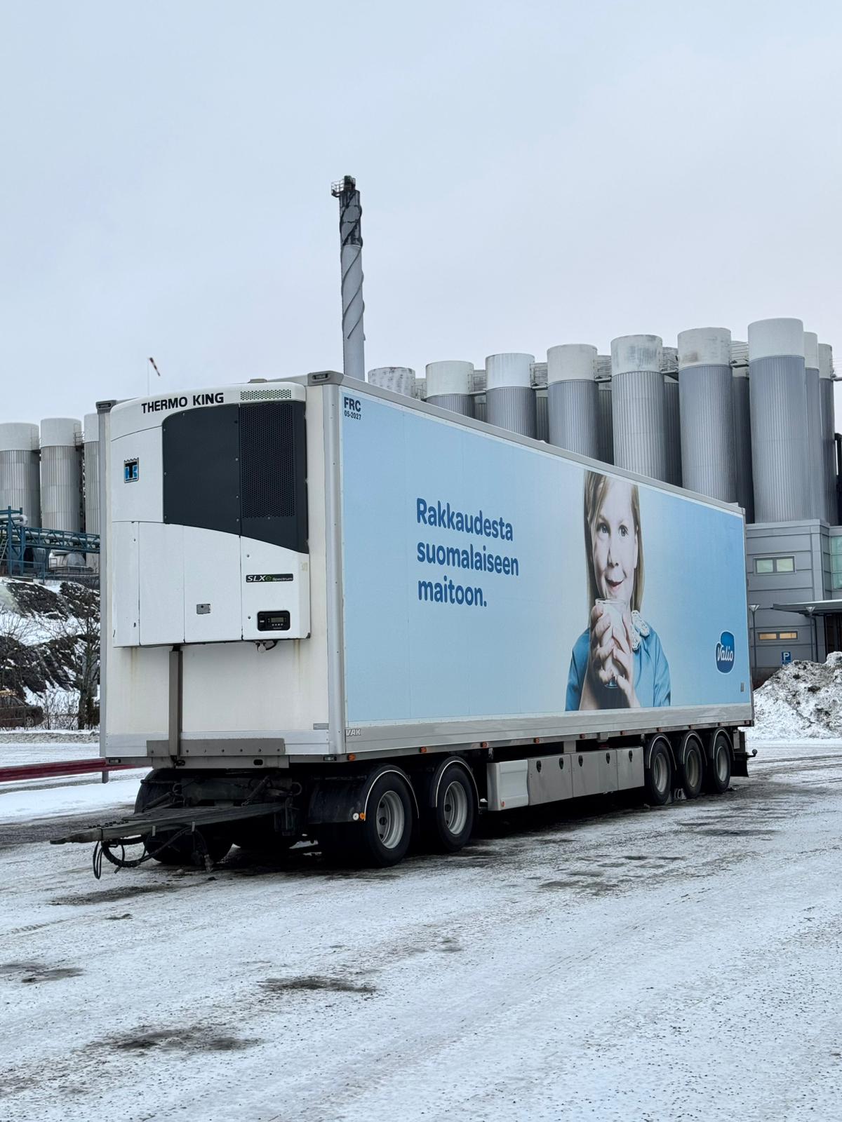 Suuri kuorma-auton peräkärry, jossa on jäähdytysyksikkö, on pysäköity lumiselle maalle. Traileri sisältää mainoksen, jossa nainen pitelee maitoa. Sivulla näkyy muulla kuin englanninkielisellä tekstillä. Tehdassiilot ovat taustalla.