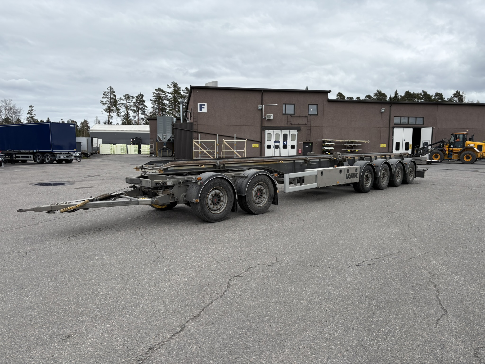 Pitkä, moniakselinen kuorma-auton perävaunu on pysäköitynä asfalttialueelle ruskean teollisuusrakennuksen eteen pilvisen taivaan alla. Taustalla näkyy puita ja muita ajoneuvoja.