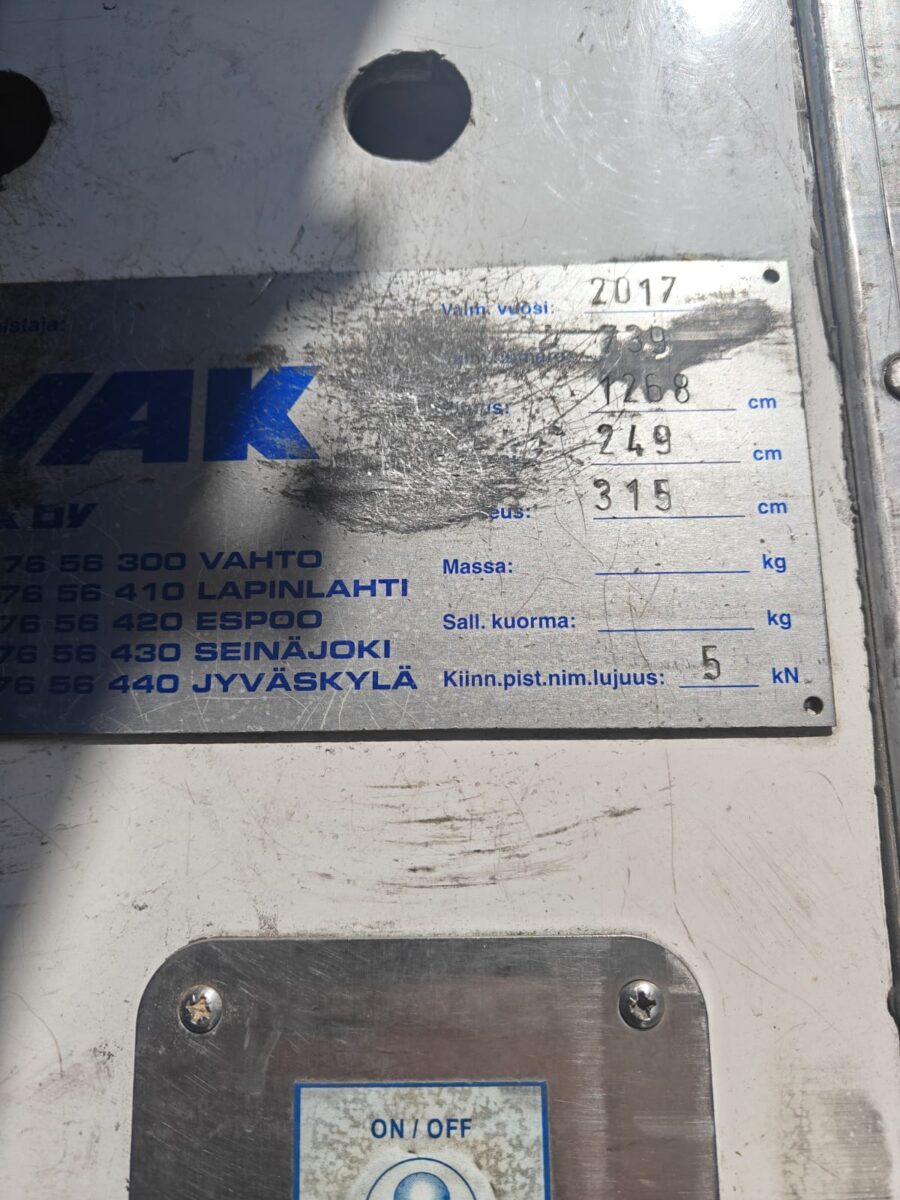 Koneen metallisessa tunnistekilvessä on tietoja, kuten vuosi (2017), tyyppi (748) ja painot (249 kg, 315 kg), ja osa tekstistä on tahrojen peitossa. Sininen virtapainike näkyy sen alla.