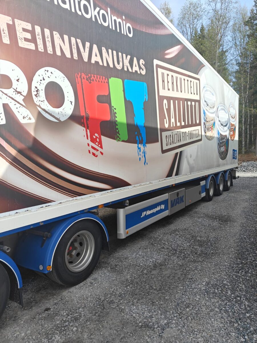 Suuri perävaunukuorma-auto, jossa on ProFit-maitotuotteiden mainoksia värikkäillä tuotekuvilla ja suomenkielisillä teksteillä, on pysäköitynä soratielle metsäalueen viereen.