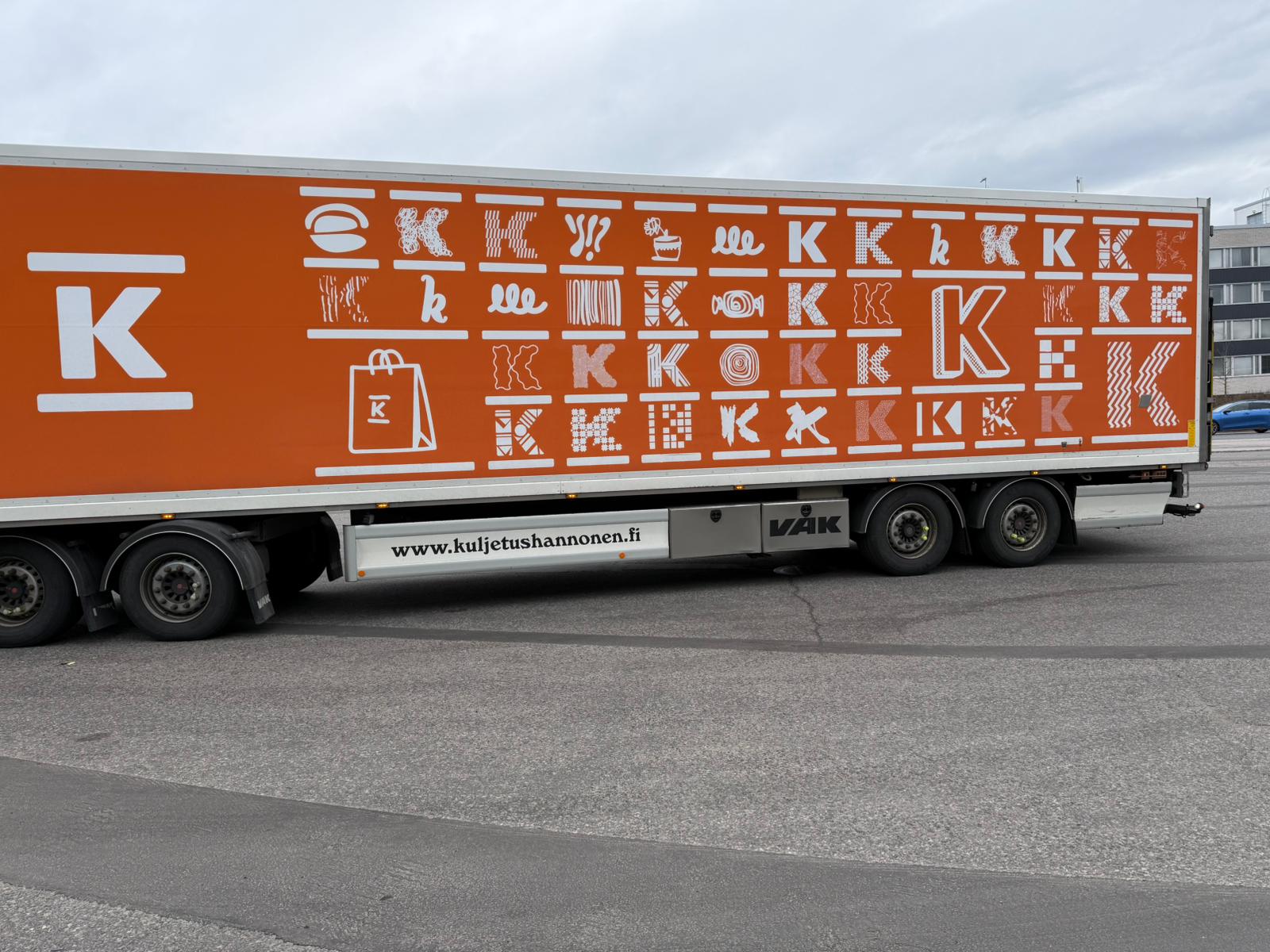 Suuressa oranssissa kuorma-auton perävaunussa on lukuisia valkoisia tyyliteltyjä K-logoja ja shoppailuun liittyviä kuvakkeita, kuten laukkuja ja kärryjä. Sivusto www.kuljetushannonen.fi näkyy kyljessä.