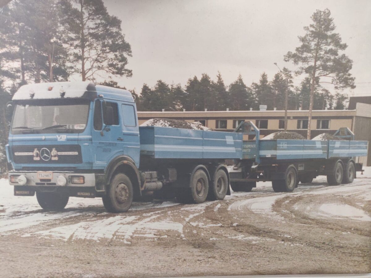 Sininen Mercedes-Benz-kippiauto perävaunuineen on pysäköitynä lumiselle, sorapäällysteiselle maalle rakennuksen ja puiden eteen sorakuormaa kuljettaen.