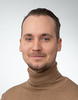 Lyhythiuksinen vaaleanruskea tukka ja siisti partainen mies hymyilee hieman. Hänellä on yllään ruskea poolopaita ja hän on poseerannut yksinkertaista, vaaleanharmaata taustaa vasten.
