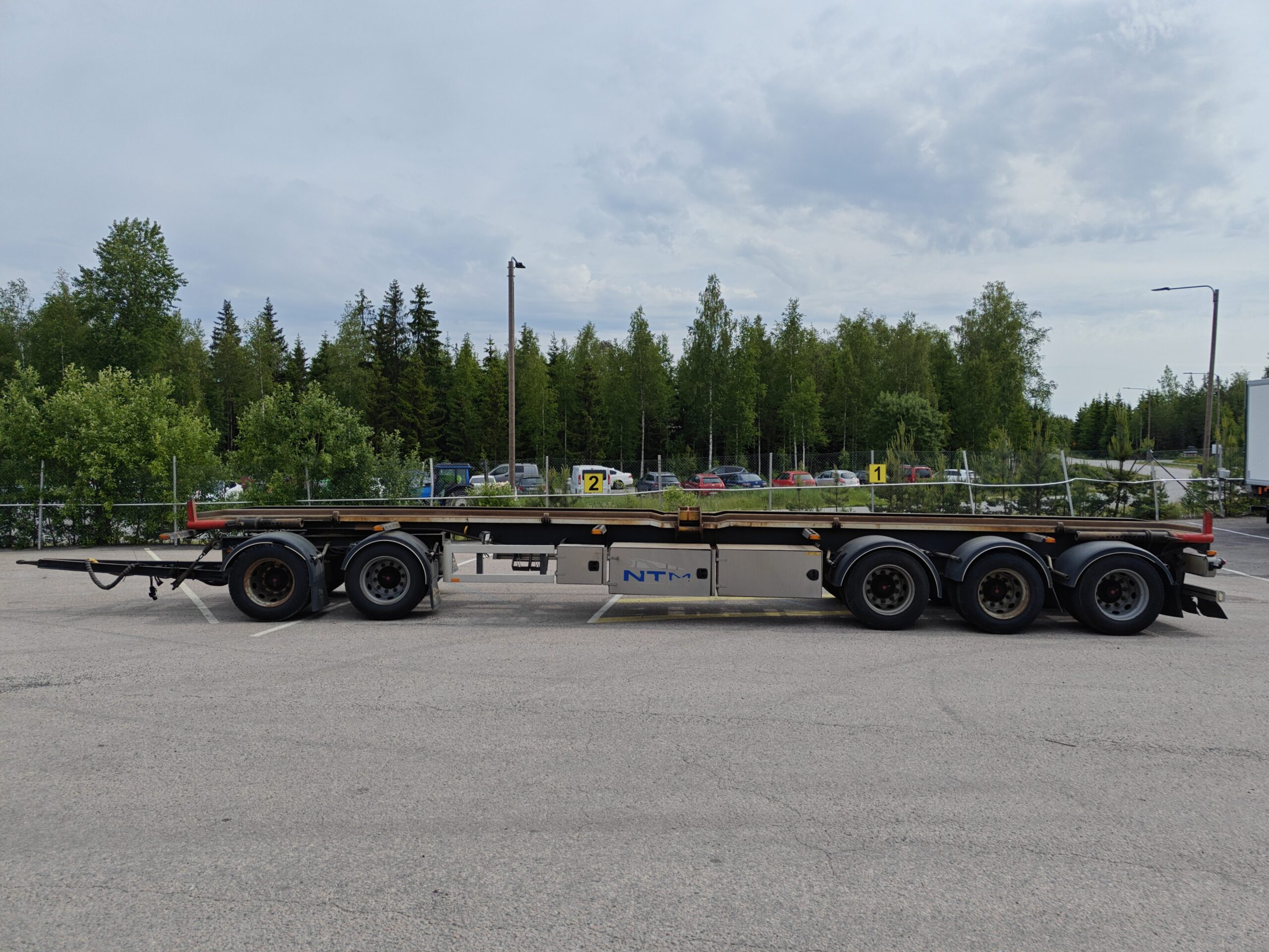 Pitkä, tyhjä, kolmiakselinen lava-autovaunu pysäköitynä asfalttipinnalle, puiden ja viherkasvien ympäröimänä pilvisen taivaan alla.