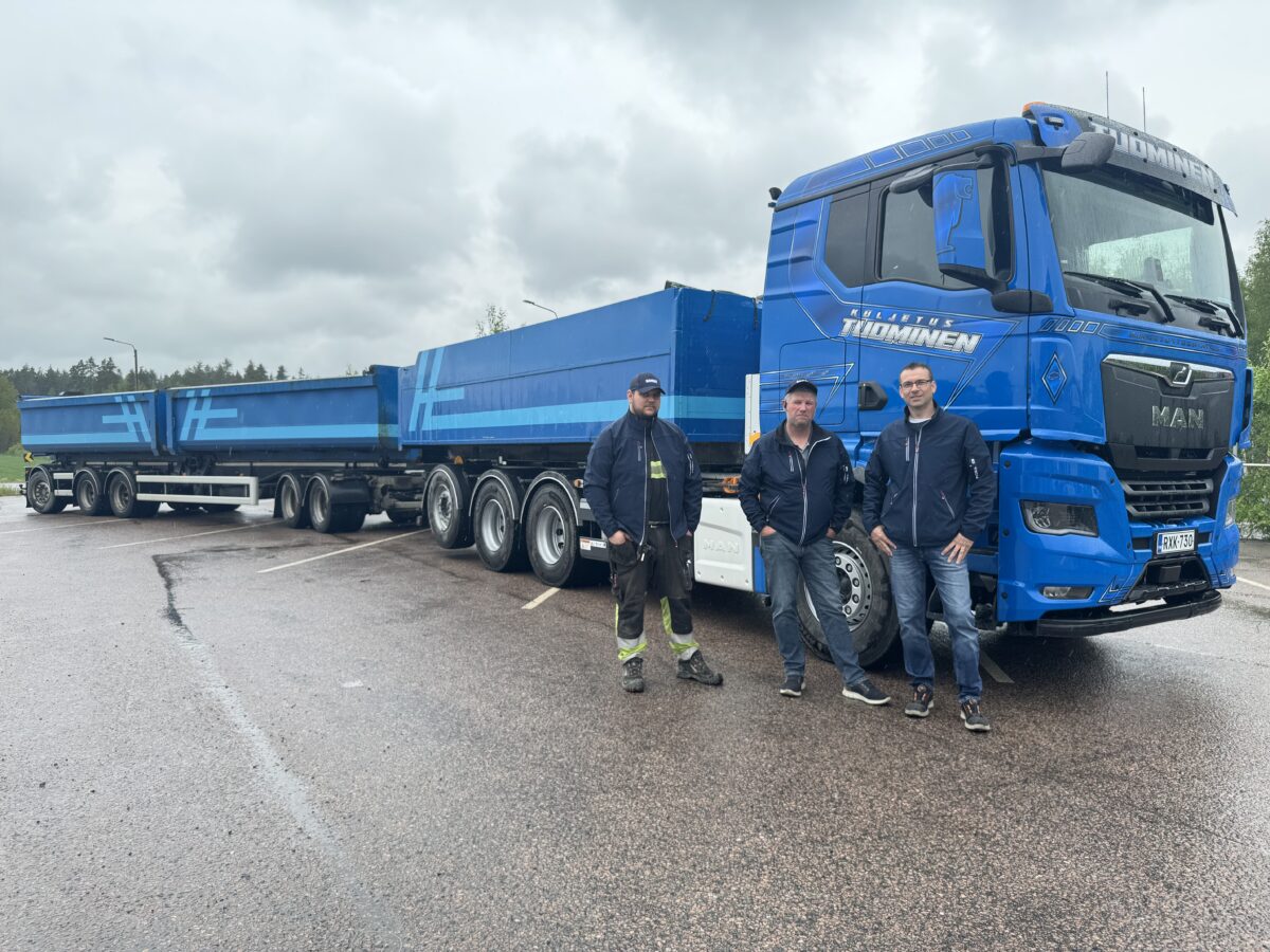 Kolme miestä seisoo suuren sinisen MAN-rekan ja samanvärisen sinisen perävaunun edessä märällä tiellä pilvisen taivaan alla. Kuorma-autossa lukee ”Tölkkö & Tömmönen”. Taustalla näkyy puita.