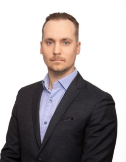 Lyhyet, vaaleanruskeat hiukset ja vaaleat silmät omaava mies, jolla on tumma bleiseri vaaleansinisen napitetun paidan päällä, poseeraa valkoista taustaa vasten.
