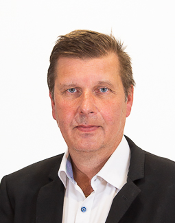 Mies jolla on lyhyet ruskeat hiukset ja joka on pukeutunut mustaan bleiseriin ja valkoiseen kauluspaitaan, poseeraa yksinkertaista valkoista taustaa vasten.