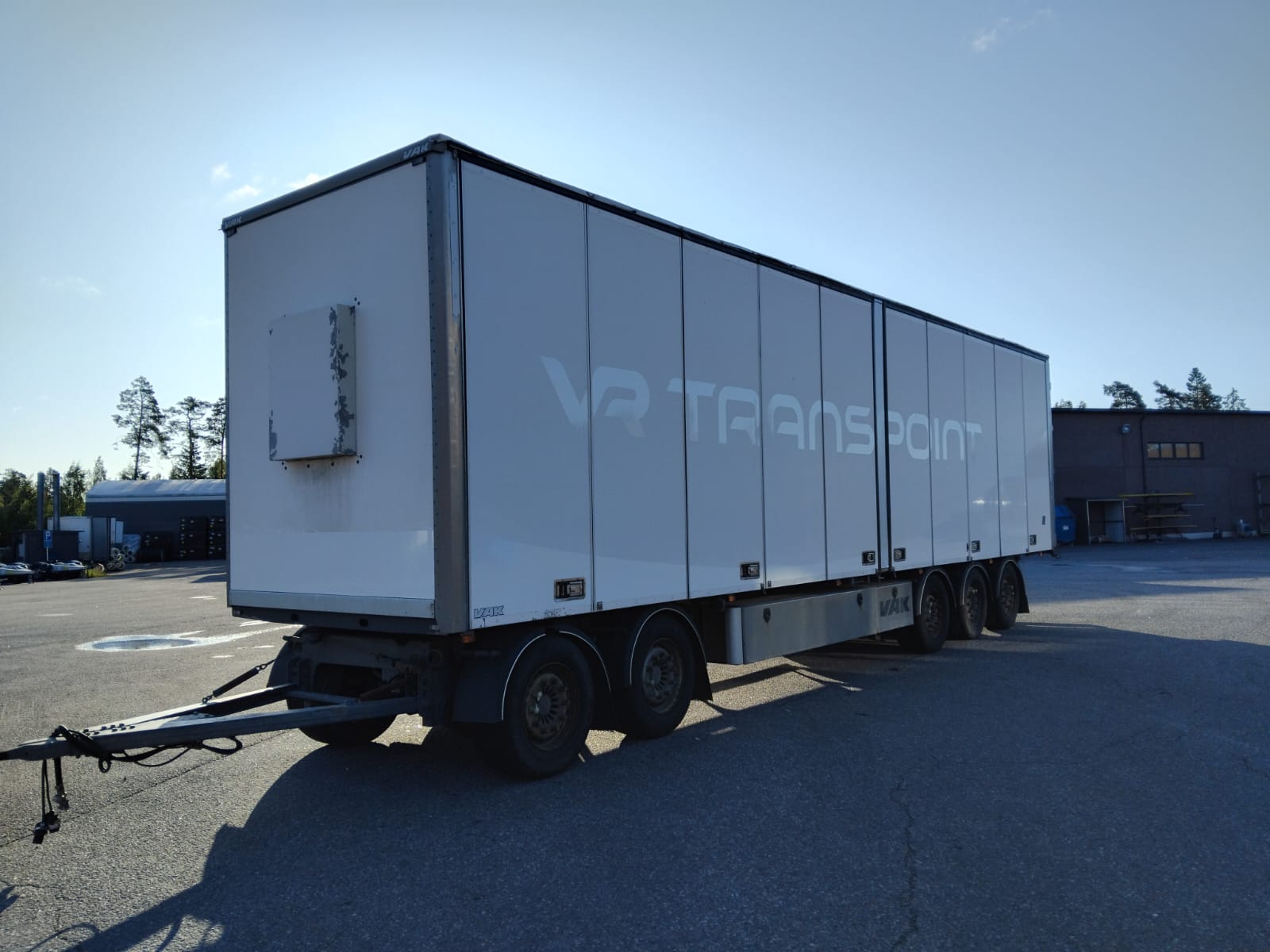 Suuri valkoinen perävaunu, johon on merkitty VR TRANSPORT, on pysäköity ulkotontille kirkkaan sinisen taivaan alla, ja taustalla näkyy joitakin rakennuksia ja puita.