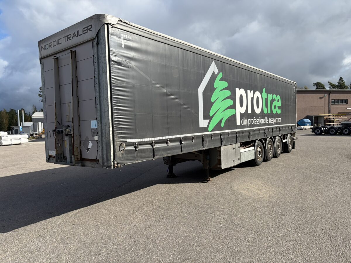 Suuri harmaa pohjoismainen perävaunu, jonka kyljessä luki ”protree din professionelle træpartner”, oli pysäköity asfalttipihalle teollisuusrakennusten lähelle.