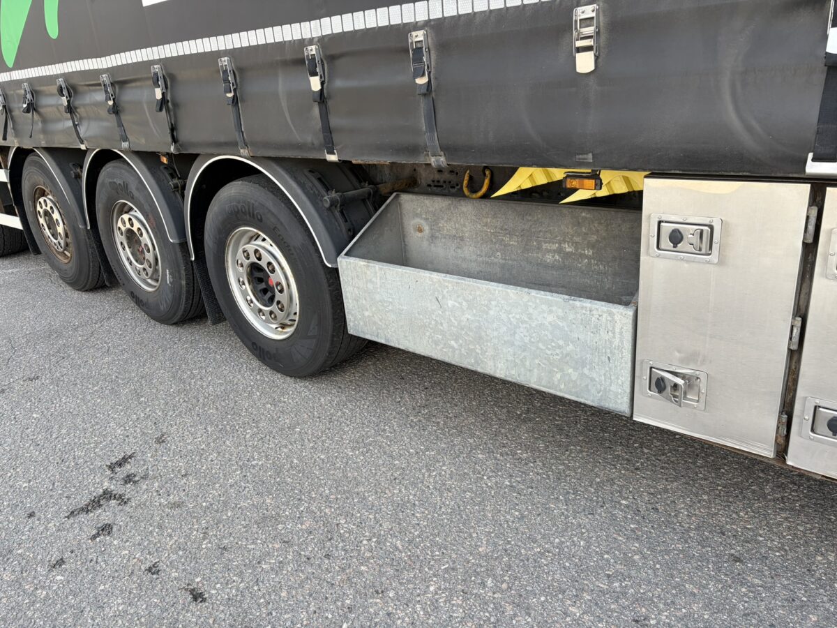 Läheltä kuvatussa kuorma-auton sivukuvassa näkyy kolme pyörää, metallinen säilytyslokero, jonka kansi on auki, sekä musta pressu, joka peittää yläosan. Kuorma-auto on pysäköity päällystetylle pinnalle.