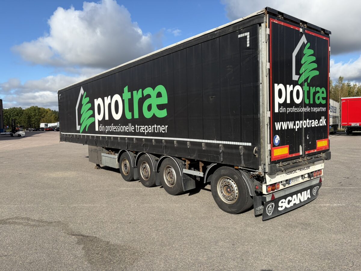 Suuri musta perävaunu, jossa on protræ-logo, on pysäköity asfalttipäällysteelle osittain pilvisen taivaan alla. Perävaunussa on kuusi pyörää, ja sen kylkeen on painettu verkkosivusto protrae.dk.