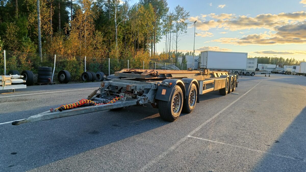 Monipyöräinen lava-perävaunu on pysäköity ulkoilma-alueelle renkaiden ja puiden kasojen viereen osittain pilvisen taivaan alla auringonlaskun aikaan.