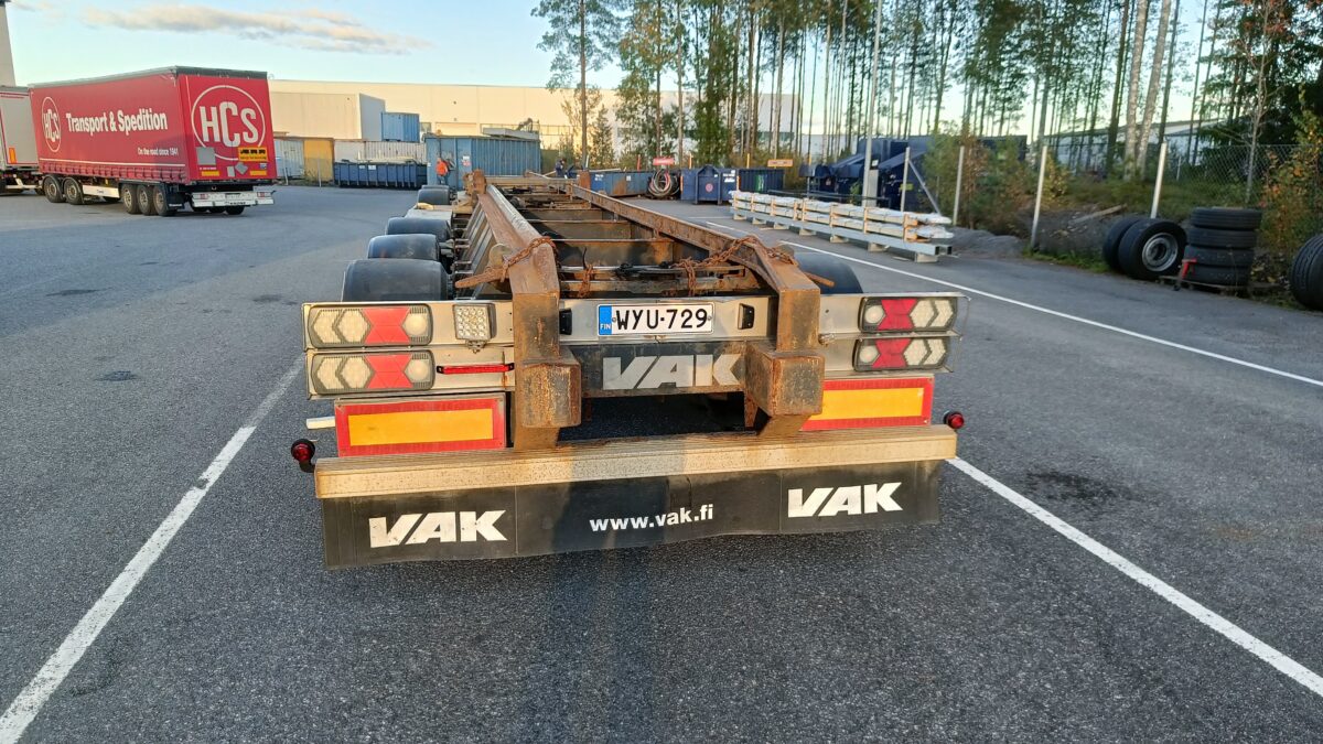 Ruosteinen kuorma-auton perävaunu, jonka rekisterinumero on HYU-729, on pysäköity puiden ja teollisuusrakennusten lähellä sijaitsevalle pysäköintialueelle. Perävaunun lokasuojassa lukee ”VAK”, ja se on tyhjä.