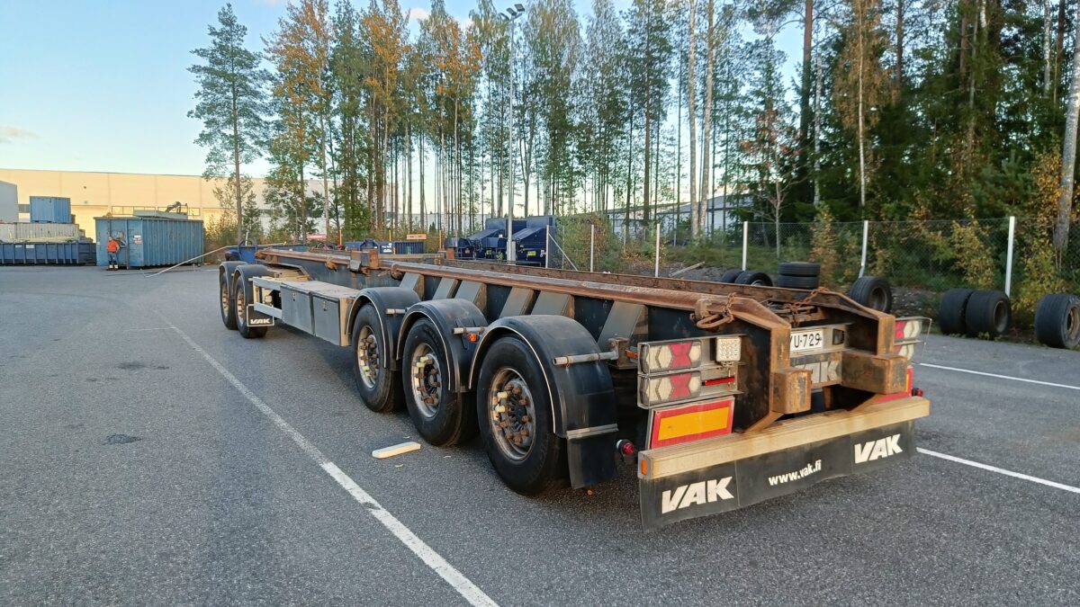 Pitkä, tyhjä kolmiakselinen perävaunu on pysäköity teollisuusalueelle lähellä metalliverkkoaitaa, metsää ja hajallaan olevia renkaita. Paikkaa valaisee myöhäisen iltapäivän auringonvalo.