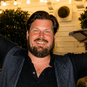 Hymyilevä mies, jolla on parta ja tummat, aaltoilevat hiukset, seisoo ulkona yöllä ja nostaa kätensä ylös. Hämärässä taustassa näkyy lämpimiä valoja ja vehreyttä.