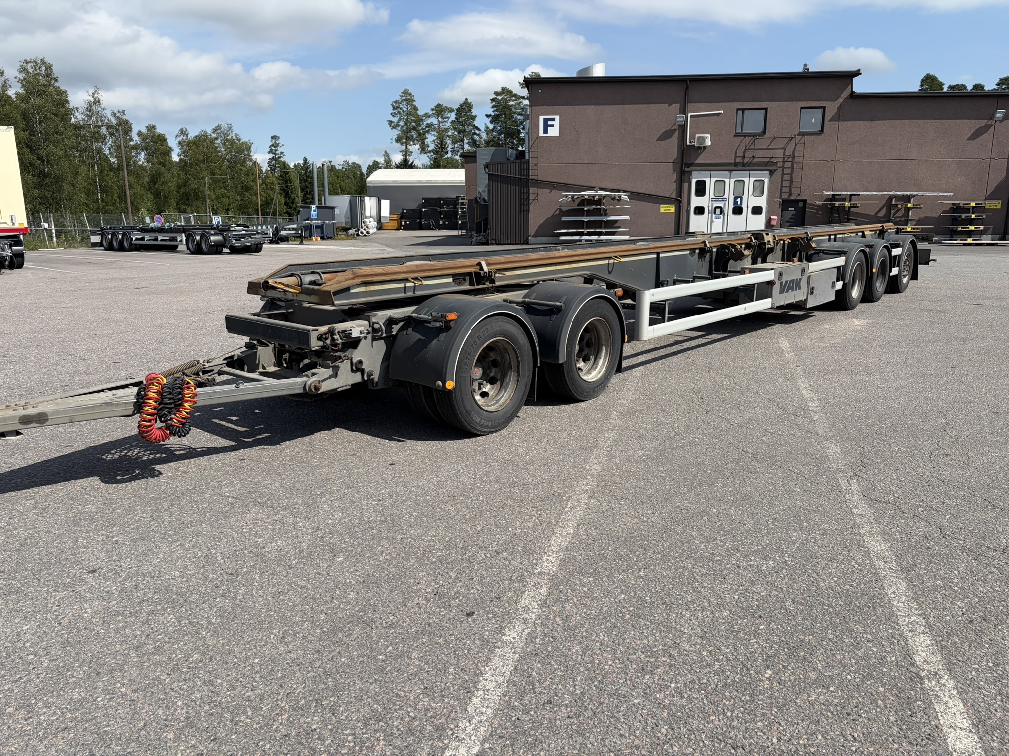 Pitkä, tyhjä kolmiakselinen puoliperävaunun alusta on pysäköity asfaltoidulle pihalle teollisuusrakennuksen ulkopuolelle osittain pilvisen taivaan alla.