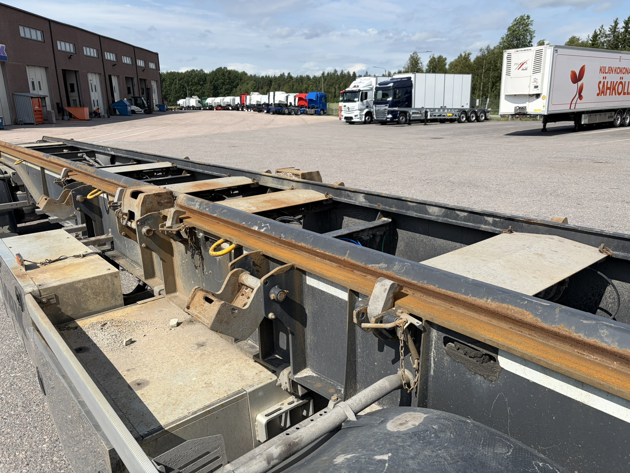 Lähikuva ruosteisesta metallikiskosta kuorma-auton alustalla pysäköintialueella. Taustalla on useita kuorma-autoja ja perävaunuja pysäköitynä rakennuksen lähellä; kuvassa näkyy puita ja osittain pilvinen taivas.