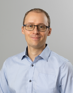 Silmälaseja ja vaaleansinistä napituspaitaa käyttävä henkilö hymyilee hieman kameraan päin, yksinkertaista vaaleanharmaata taustaa vasten.
