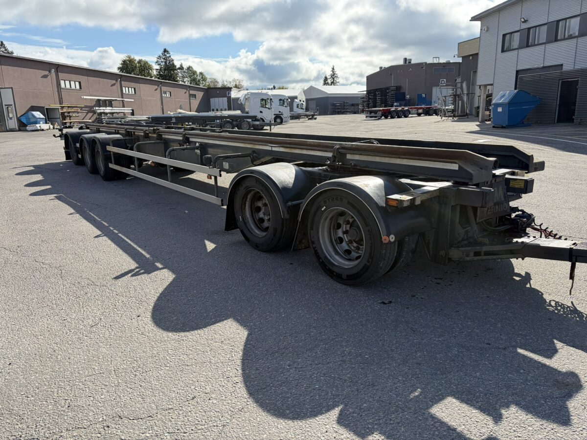 Pitkä, tyhjä moniakselinen lava-perävaunu on pysäköity teollisuusalueelle useiden rakennusten lähelle osittain pilvisen taivaan alla.
