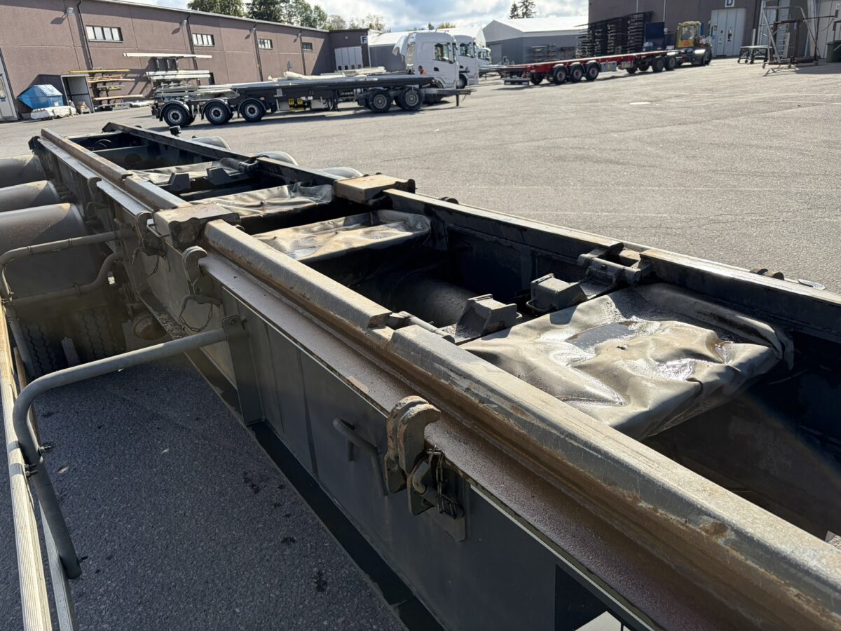 Lähikuva metallisesta kuorma-auton alustasta teollisuusalueella; taustalla näkyy muita kuorma-autoja ja perävaunuja kirkkaan taivaan alla.