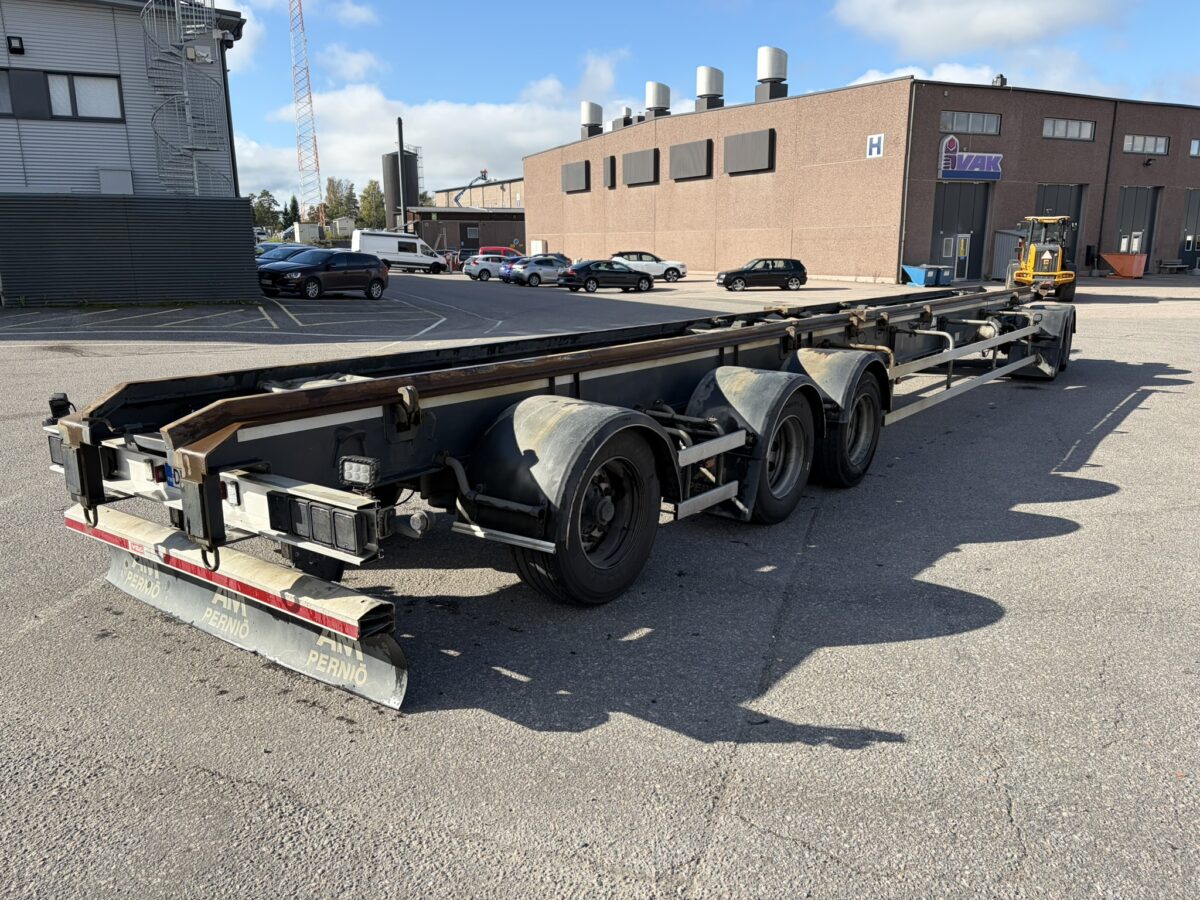 Pitkä, tyhjä kolmiakselinen perävaunu on pysäköity asfalttipihalle teollisuusrakennusten ja ajoneuvojen viereen osittain pilvisen taivaan alla.