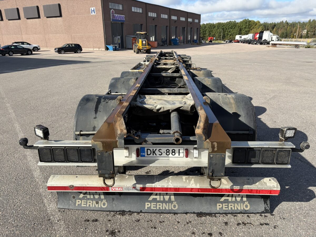 Lähikuva tyhjän kuorma-auton alustan takaosasta pysäköintialueella; kuvassa näkyy rekisterikilpi DKS-881 ja lokasuojat, joissa lukee ”AM PERNIÖ”. Taustalla näkyy teollisuusrakennuksia ja ajoneuvoja.