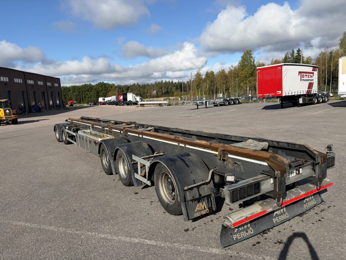 Monipyöräinen lava-perävaunu on pysäköity asfalttipäällysteelle teollisuusrakennusten ja puiden lähelle osittain pilvisen taivaan alla.