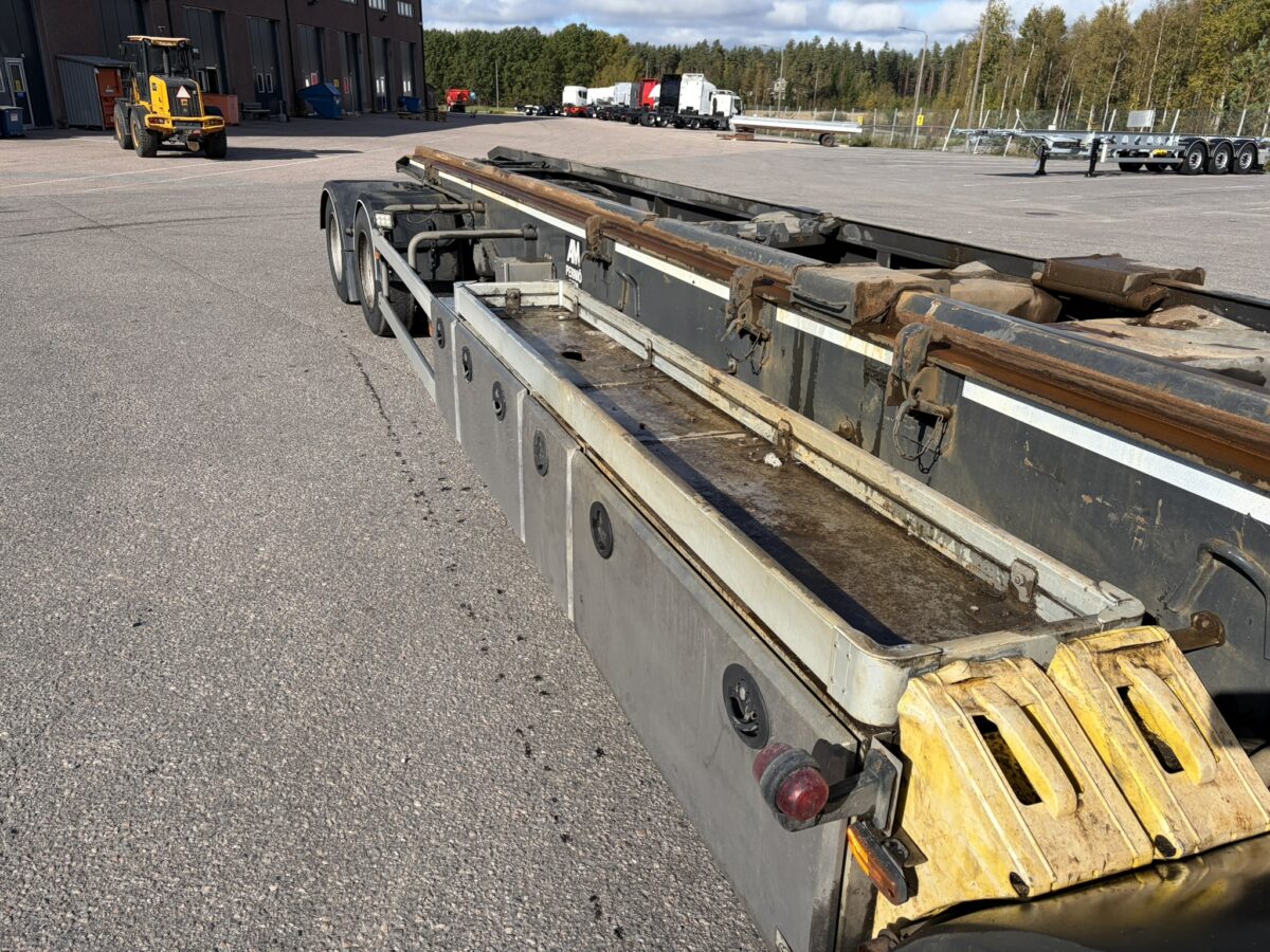 Lähikuva sään kuluttamasta kuorma-auton perävaunun alustasta, joka on pysäköity asfalttipäällysteelle. Taustalla näkyy puita, perävaunuja ja keltainen rakennustyöajoneuvo kirkkaan taivaan alla.