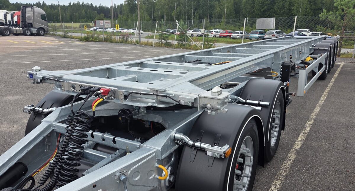 Lähikuva moniakselisesta metallisesta kuorma-auton alustasta, joka on pysäköity asfalttipäällysteelle aidatun alueen läheisyyteen; taustalla näkyy autoja ja metsä.