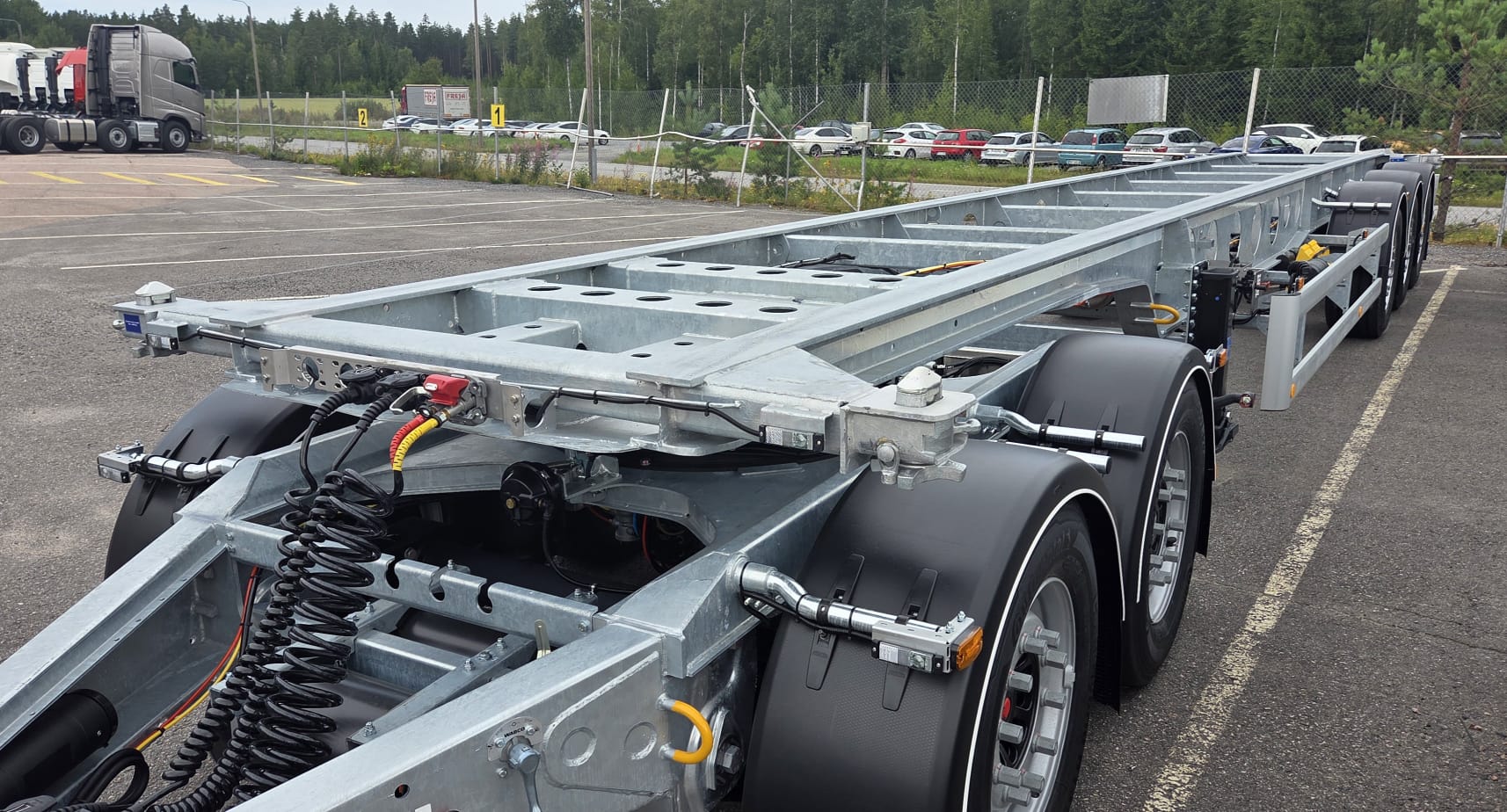 Lähikuva moniakselisesta metallisesta kuorma-auton alustasta, joka on pysäköity asfalttipäällysteelle aidatun alueen läheisyyteen; taustalla näkyy autoja ja metsä.
