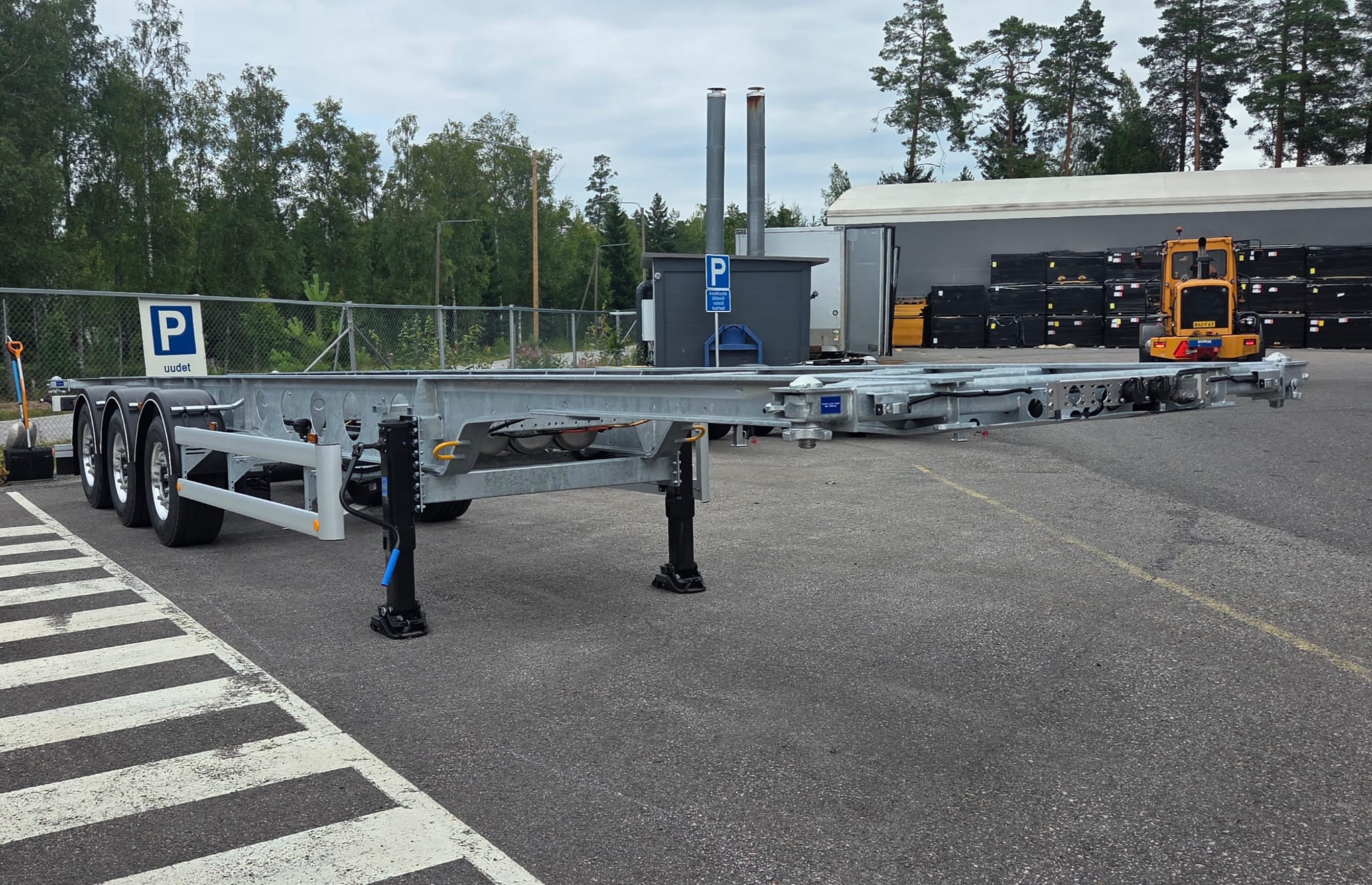 Kolmiakselinen metallinen perävaunun runko on pysäköity tontille aidan, puiden ja rakennuksen läheisyyteen. Taustalla näkyy pieni rakennustyömaajoneuvo ja pysäköintikyltejä.