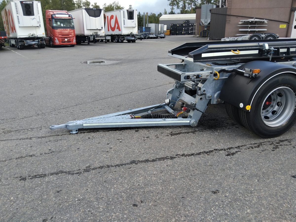 Lähikuva perävaunun vetokoukusta asfalttipihalla; taustalla on useita kuorma-autoja ja perävaunuja pysäköitynä rakennuksen lähellä.
