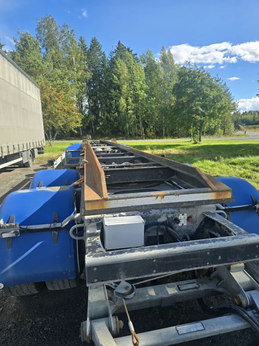 Lähikuva tasoperävaunun metallirungosta, jossa on sinisiä yksityiskohtia; vaunu on pysäköity ulos aurinkoisena päivänä, ja sitä ympäröi vihreä ruoho ja puut.