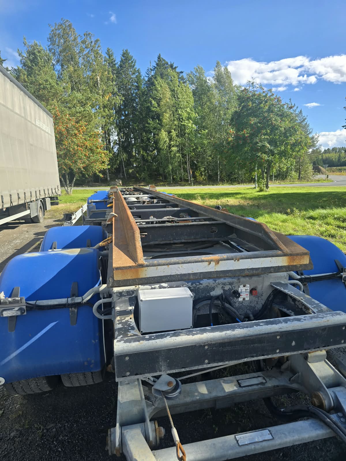 Lähikuva tasoperävaunun metallirungosta, jossa on sinisiä yksityiskohtia; vaunu on pysäköity ulos aurinkoisena päivänä, ja sitä ympäröi vihreä ruoho ja puut.