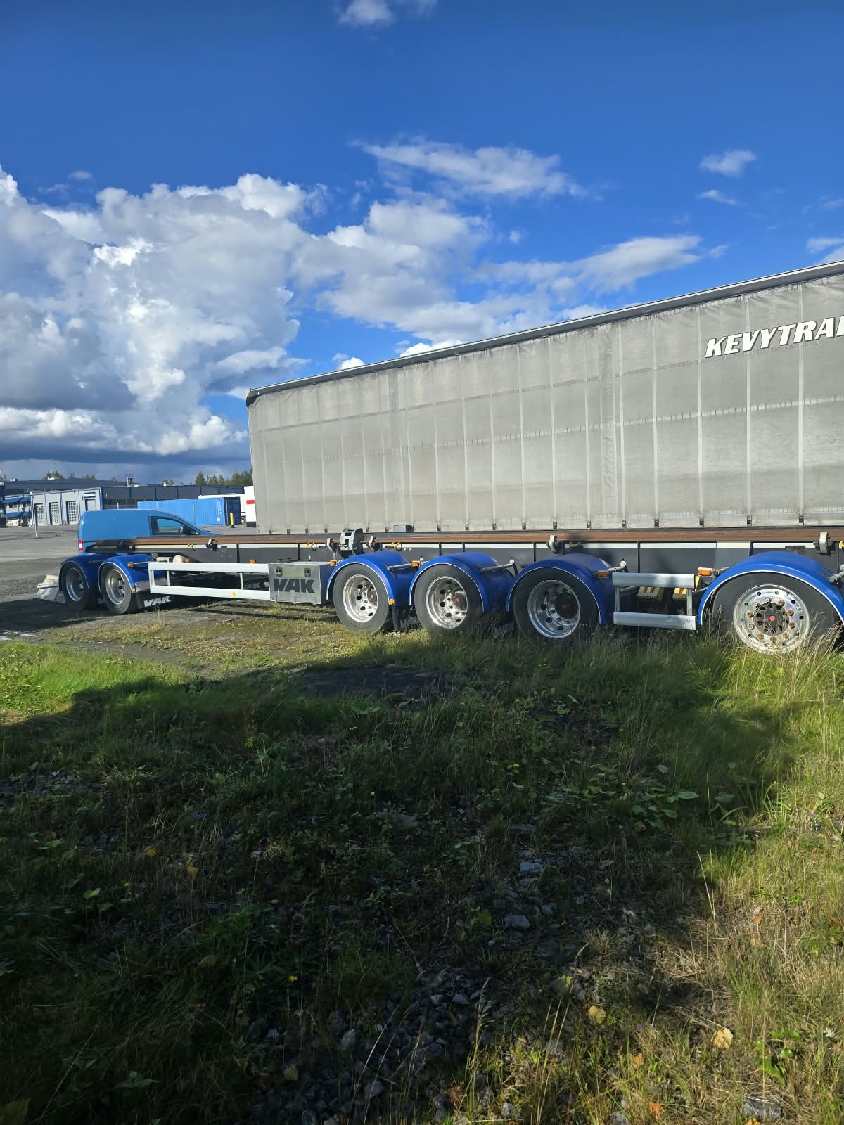 Sininen puoliperävaunukuorma-auto, jossa on katettu perävaunu, on pysäköity soralle nurmikon ääreen kirkkaan sinisen taivaan alla, jossa on hajanaisia pilviä.