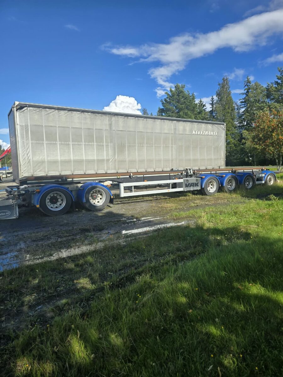 Suuri, harmaa lava-perävaunu on pysäköity nurmikolle puiden lähelle sinisen, pilvisen taivaan alla. Perävaunussa on kolme akselia ja siniset vanteet.