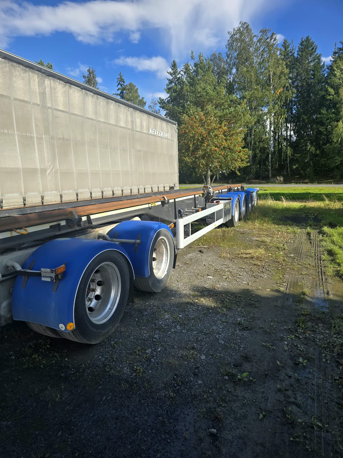 Sinivalkoinen puoliperävaunu on pysäköity soratielle nurmialueen läheisyyteen, ja sitä ympäröivät vihreät puut osittain pilvisen taivaan alla.