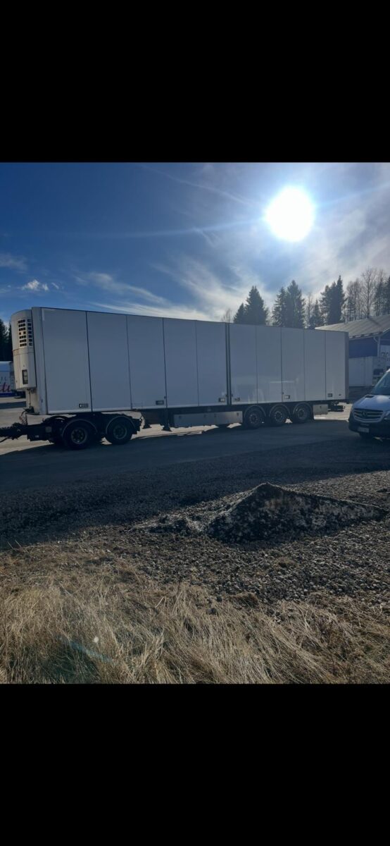 Suuri valkoinen kuorma-auton perävaunu on pysäköity asfalttitielle kirkkaan auringon alla, ja taustalla näkyy puita ja rakennuksia. Perävaunun vieressä näkyy osittain auto, ja etualalla on nurmikkoalue.