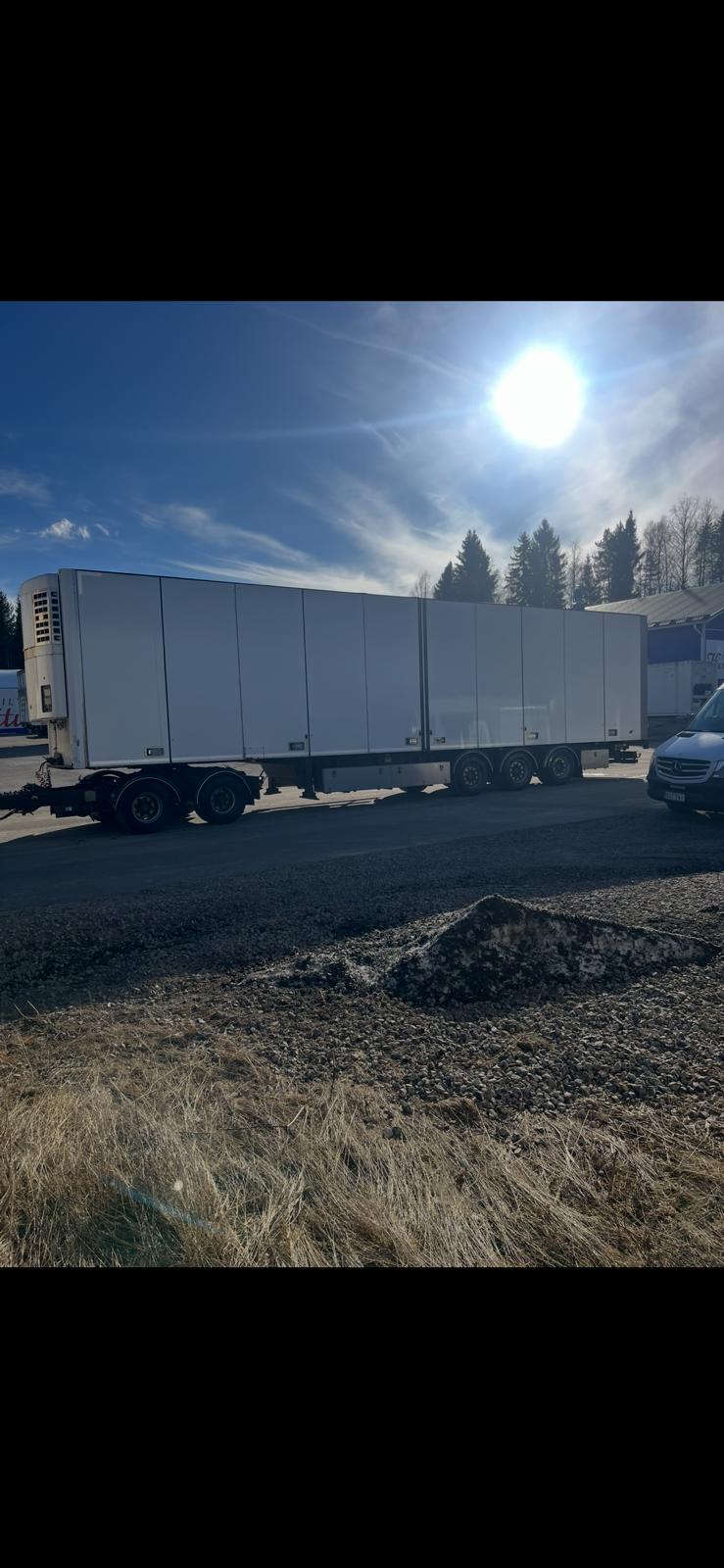 Suuri valkoinen kuorma-auton perävaunu on pysäköity asfalttitielle kirkkaan auringon alla, ja taustalla näkyy puita ja rakennuksia. Perävaunun vieressä näkyy osittain auto, ja etualalla on nurmikkoalue.